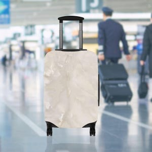 May include: A cream-colored rolling suitcase with a marble-like pattern. The luggage has a black handle and wheels, and is in an airport setting. The suitcase is upright and ready for travel.