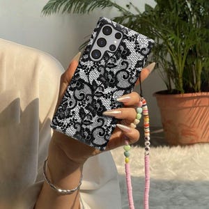 May include: A rectangular smartphone case featuring a detailed black lace pattern on a white background. The case has a cutout for a multi-lens camera system. Attached to the side is a beaded phone strap with colorful discs and letter beads.
