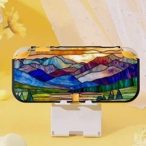 以下が含まれることがあります： 青、紫、緑、オレンジの色合いで、ステンドグラス風の山々が描かれた携帯ゲーム機のケース。ケースには黄色のアクセントがあり、白いスタンドに置かれています。風景を連想させるデザインです。