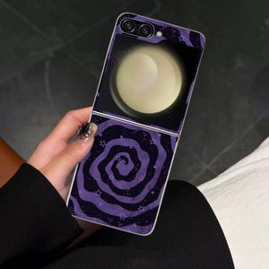 Puede incluir: Una funda para smartphone plegable con un diseño en tonos morado oscuro y negro. La sección superior muestra una gran luna de color crema brillante sobre un fondo oscuro, mientras que la inferior presenta un patrón de espiral morada con pequeñas estrellas.