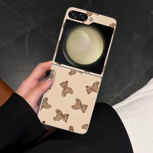 May include: A beige mobile phone case featuring a repeating pattern of brown teddy bear illustrations. The phone's screen shows a bright, circular image. The case is held in a person's hand.