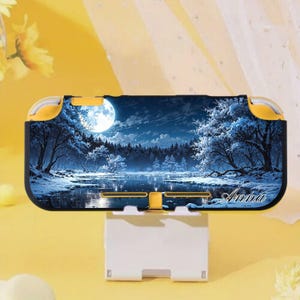 Puede incluir: Una funda amarilla y negra para Nintendo Switch Lite con un paisaje invernal, una luna llena, árboles y un lago. El nombre "Anna" está escrito en cursiva en la parte inferior derecha. La funda está sobre un soporte blanco.