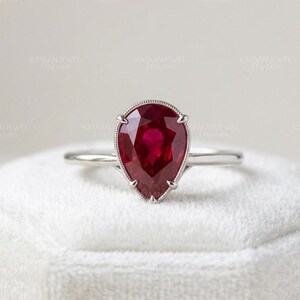 Puede incluir: Un anillo de plata con una gran gema roja oscura en forma de pera. La gema está engastada en un bisel delicado y detallado. El anillo se exhibe sobre una superficie blanca y texturizada. Es una joya.