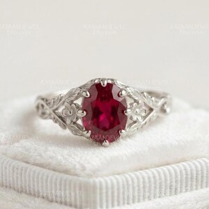 Pode incluir: Um anel de tom prateado com uma pedra preciosa de cor vermelho-profundo de corte oval no centro. A banda apresenta um trabalho de metal intrincado que retrata pequenas flores e folhas que se estendem da pedra central, criando um design botânico.