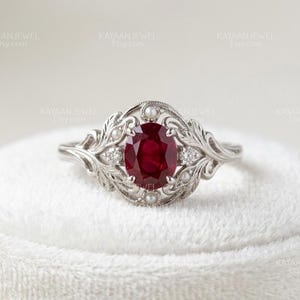May include: A silver-toned ring featuring a central oval-cut deep red ruby, flanked by two small round diamonds and accented with tiny white pearls. The metal band displays intricate leaf-like filigree patterns surrounding the gemstones.