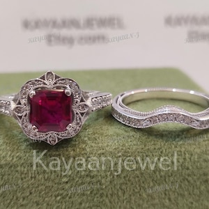 Puede incluir: Un juego de anillos de dos piezas con una piedra preciosa roja cuadrada en un engaste de halo. El anillo de compromiso tiene detalles intrincados y se combina con una banda curva, ambos en plata.