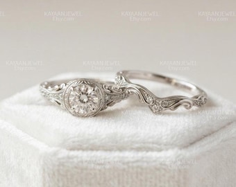 May include: A silver-toned engagement ring set featuring a round center stone surrounded by intricate milgrain detailing and scrollwork, paired with a matching curved wedding band adorned with small accent stones along the band's ornate metalwork.