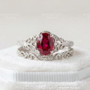 Könnte beinhalten: Ein Ring-Set in Silberoptik mit einem oval geschliffenen, tiefroten Edelstein als Herzstück. Der Ring und das passende, geschwungene Ehering-Band sind mit filigranen Metallarbeiten und kleinen, klaren Steinen an den Seiten verziert.