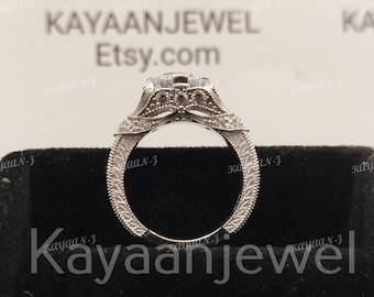 Op de afbeelding: Een zilveren verlovingsring met een grote, heldere middelste steen en kleinere stenen die zijn gezet in een delicaat, ingewikkeld ontwerp. De ring wordt getoond op een zwarte fluwelen doos met de tekst "Kayaanjewel" gedrukt op de voorkant.