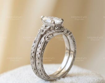 May include: A silver-toned engagement ring set featuring a marquise-cut center stone held by prongs. The band and matching wedding band are crafted with intricate leaf-like engravings and small embedded stones along the sides of the metal shank.
