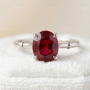 Puede incluir: Un anillo de plata con una gran piedra preciosa de color rojo intenso y corte ovalado, sujeta por cuatro garras. El aro del anillo tiene un acabado liso y pulido con un diseño segmentado sutil en cada lado, creando una apariencia limpia y refinada.