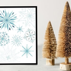 Può includere: Una stampa d'arte stampabile con un design ad acquerello di fiocchi di neve blu su sfondo bianco. La stampa è incorniciata in una cornice nera ed è esposta su una superficie bianca.