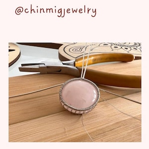 May include: A close-up of a pink gemstone pendant being wrapped in silver wire. The pendant is being worked on with pliers on a wooden surface.