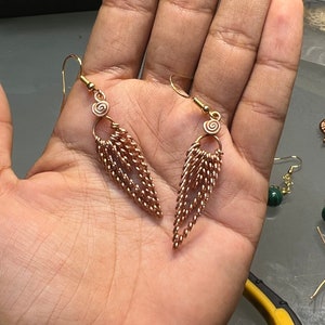 Peut inclure: Une paire de boucles d'oreilles dorées avec un motif en spirale en haut et une chaîne en cascade de petites boucles de fil torsadé. Les boucles d'oreilles pendent de la main d'une personne.