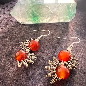 May include: A pair of dangle earrings featuring large, round, orange-red beads. The beads are accented with silver-colored metal filigree and hooks. The earrings are displayed against a dark, textured background.