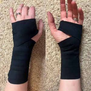 May include: Two hands with black wrist wraps. The wraps are tightly wound around the wrists and partially cover the hands. The wraps are made of a soft, flexible material.