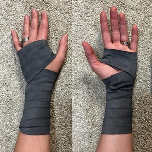 May include: A dark gray wrist wrap made of a soft, flexible fabric that covers the forearm and extends to the base of the fingers. The wrap features a thumb hole and is secured with multiple overlapping layers of material around the wrist area.