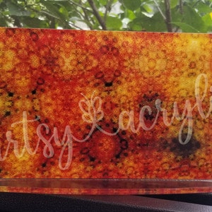 May include: A rectangular piece of acrylic with a red, orange, and yellow abstract pattern. The acrylic is on a clear acrylic stand. The text "artsy acrylic" is written on the acrylic.