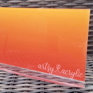 May include: A rectangular acrylic sheet with a gradient of orange to yellow, displayed in a clear acrylic stand. The text "artsy acrylic" is printed in white on the front of the sheet. The stand is on a woven surface.