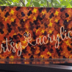 May include: A clear acrylic stand with a colorful abstract design featuring orange, yellow, and black colors. The design has a watercolor effect and the text "artsy acrylic" is written in white on the front of the stand.