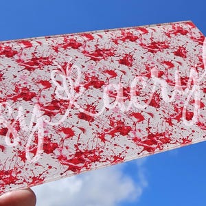 May include: A sheet of clear acrylic with a red splatter paint design. The acrylic sheet is held in a hand against a blue sky background.