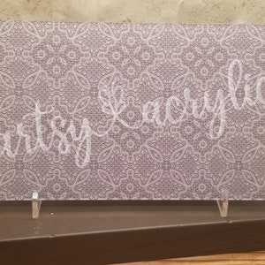 May include: A white acrylic sign with a delicate, intricate, gray and white patterned design. The sign is displayed on a brown stand with two clear acrylic supports.