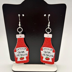 May include: A pair of novelty earrings shaped like ketchup bottles. The red glittery bottles have white labels with the text "Catch Up With Jesus" and a tomato illustration. The earrings hang from silver-colored hooks.