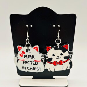 May include: A pair of cat-shaped earrings with silver hooks. One earring is white with red accents and the text "BE PURR FECTED IN CHRIST." The other is black and white with a red bow tie. The earrings are displayed on a black stand.