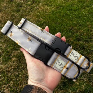 May include: Two gray dog collars with a bee and daisy pattern. The collars have black plastic buckles and metal D-rings. The text "Pup dog" is printed on a white label on each collar.