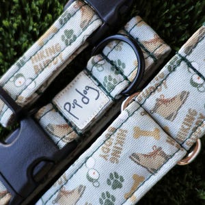 May include: A white and green dog collar with a brown and white print of hiking boots, paw prints, and bones. The collar has a black buckle and a black metal D-ring. The collar also has a white label with the text "Pup Dog" written in black.