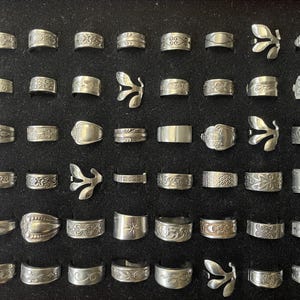 May include: A collection of silver rings displayed on a black surface. The rings feature various designs, including floral patterns, geometric shapes, and unique butterfly-shaped rings. The rings are of different widths and styles.