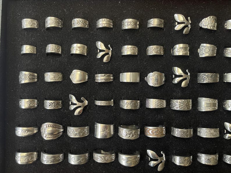 May include: A collection of silver rings displayed on a black surface. The rings feature various designs, including floral patterns, geometric shapes, and unique butterfly-shaped rings. The rings are of different widths and styles.
