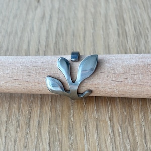 May include: A silver-colored metal ring with a leaf-like design is shown on a wooden dowel. The ring has three curved leaves and a small rectangular piece above it. The dowel is light brown and rests on a wooden surface.