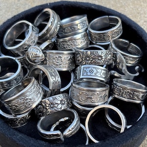 May include: A collection of silver-toned rings with floral and leaf designs, displayed in a black container. The rings vary in style, some featuring embossed patterns and others with open designs. The rings are made of metal.