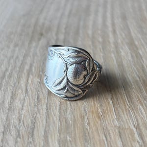 Może przedstawiać: Srebrny pierścionek ze łyżki z ozdobnym wzorem z liśćmi i centralnym, teksturowanym elementem. Pierścionek jest umieszczony na jasnej drewnianej powierzchni, prezentując swoje skomplikowane detale i estetykę vintage.