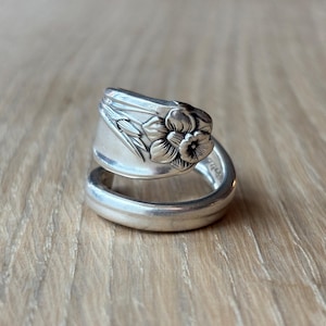 May include: A silver-toned spoon ring featuring an embossed floral design on the handle portion. The metal band wraps around to form a circular shape, showcasing detailed engravings of a flower and leaves on the curved surface of the spoon handle.