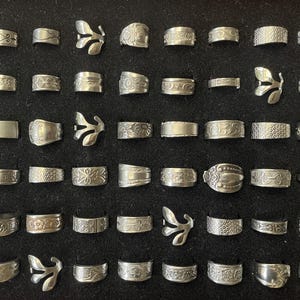 May include: A collection of silver rings displayed on a black surface. The rings feature various designs, including floral patterns, textured bands, and leaf-shaped accents. The rings are of different widths and styles.