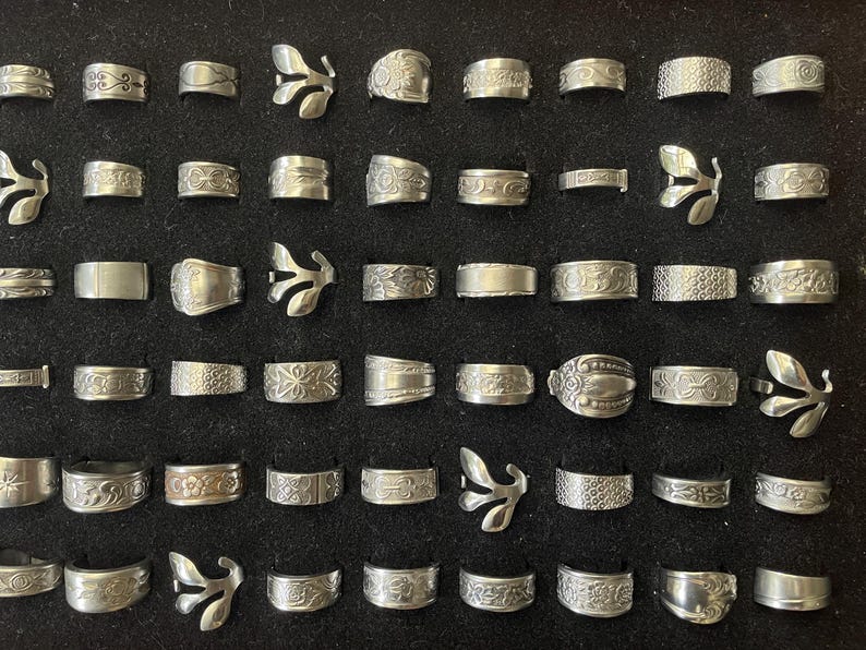 May include: A collection of silver rings displayed on a black surface. The rings feature various designs, including floral patterns, textured bands, and leaf-shaped accents. The rings are of different widths and styles.