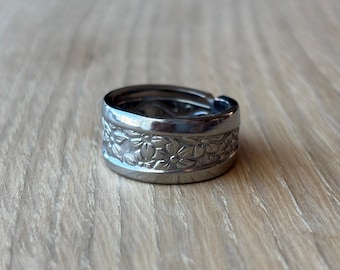 Op de afbeelding: Een zilverkleurige metalen ring met een brede band voorzien van een reliëf bloemenpatroon aan de buitenzijde. De ring heeft een gepolijste, reflecterende afwerking en een gladde, effen rand aan zowel de boven- als onderzijde.