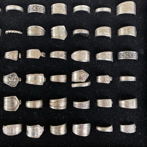 May include: A collection of silver rings with various designs, including floral patterns and textured bands. The rings are displayed on a black surface, showcasing their intricate details and craftsmanship. The rings are of different sizes and styles.