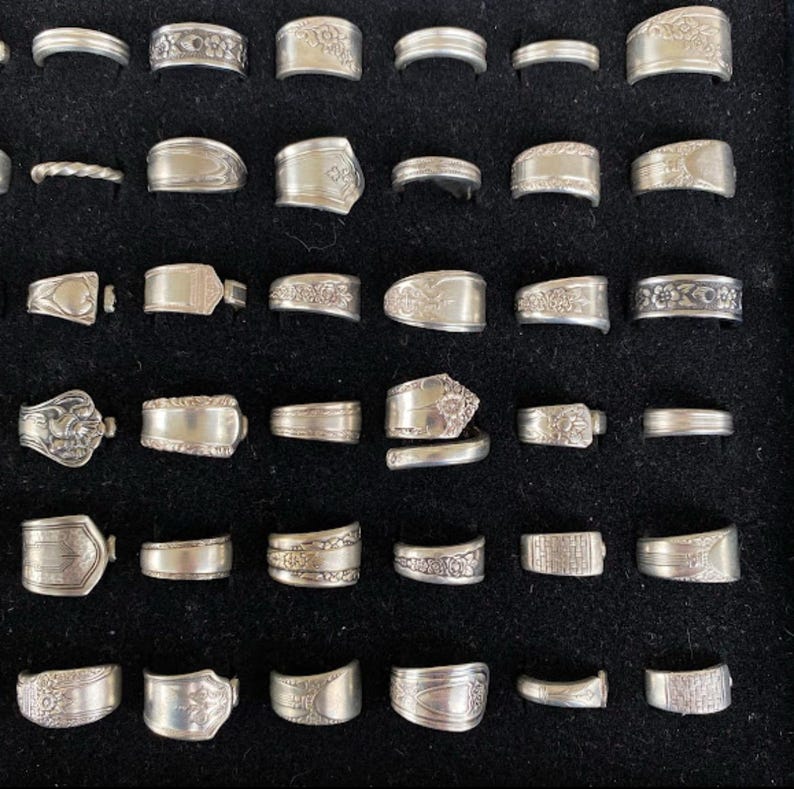 May include: A collection of silver rings with various designs, including floral patterns and textured bands. The rings are displayed on a black surface, showcasing their intricate details and craftsmanship. The rings are of different sizes and styles.
