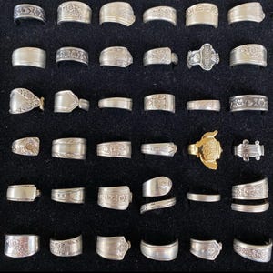 May include: Collection of silver rings with various designs, including floral patterns and engraved details. One gold-colored ring with a turtle design is also present. The rings are displayed on a black surface, showcasing their intricate craftsmanship.