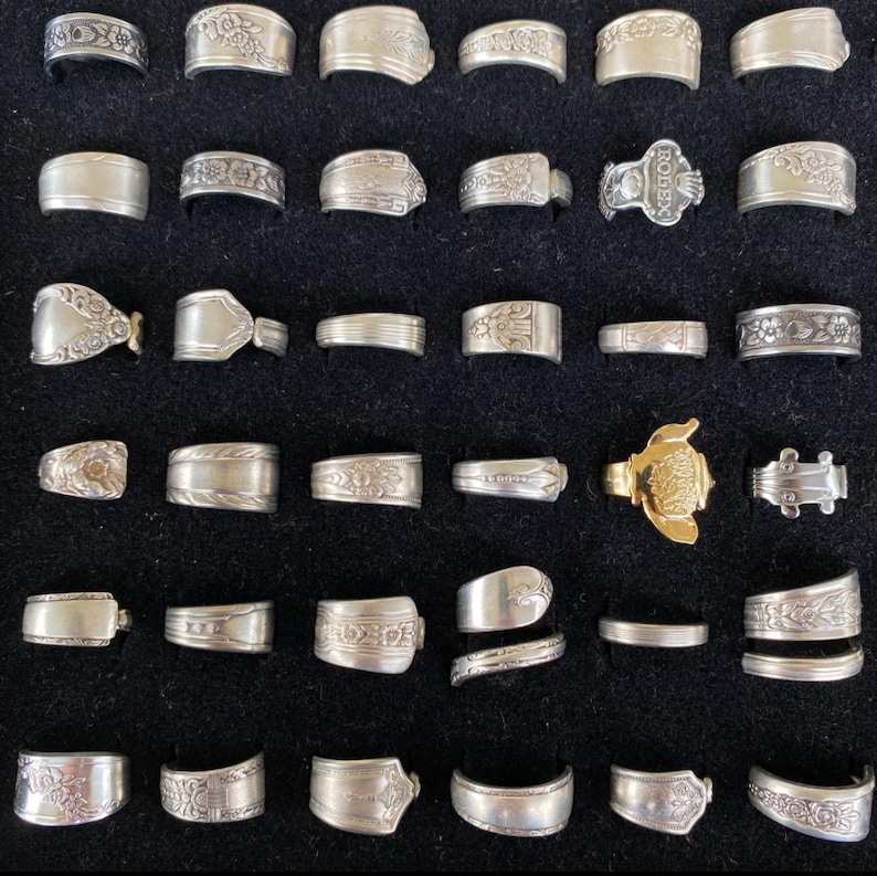 May include: Collection of silver rings with various designs, including floral patterns and engraved details. One gold-colored ring with a turtle design is also present. The rings are displayed on a black surface, showcasing their intricate craftsmanship.
