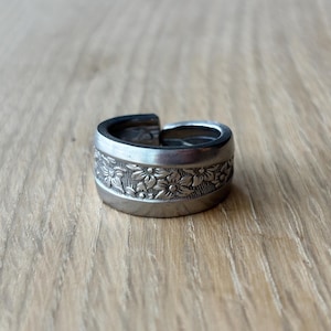 May include: A silver-toned ring with a floral design. The ring features a band of detailed flowers around the center. The ring is adjustable and sits on a wooden surface.