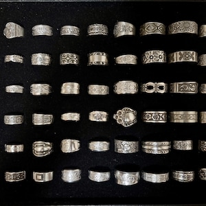 May include: A collection of silver rings with various designs, including floral, geometric, and abstract patterns. The rings are displayed on a black surface, showcasing their intricate details and textures. The rings vary in size and style.