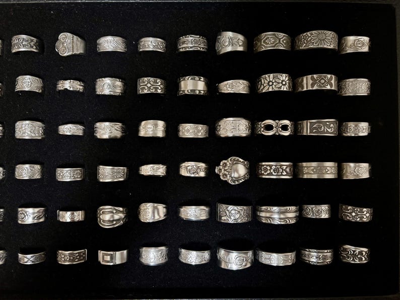 May include: A collection of silver rings with various designs, including floral, geometric, and abstract patterns. The rings are displayed on a black surface, showcasing their intricate details and textures. The rings vary in size and style.