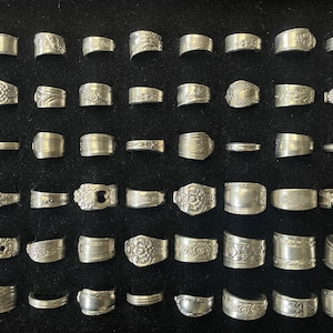 May include: Collection of silver rings with various designs, including floral, geometric, and textured patterns. The rings are displayed on a black surface, showcasing their intricate details and vintage aesthetic. The rings are made of silver.