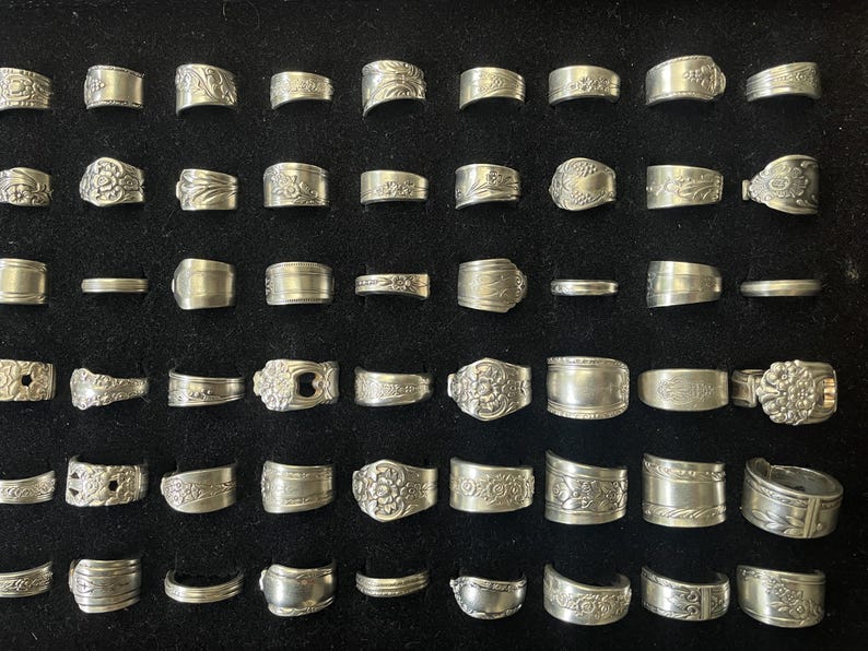May include: Collection of silver rings with various designs, including floral, geometric, and textured patterns. The rings are displayed on a black surface, showcasing their intricate details and vintage aesthetic. The rings are made of silver.