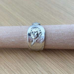 Könnte beinhalten: Ein silberner Ring mit einem Porträt von Thomas Jefferson. Der Ring, gefertigt aus einem Löffel, weist aufwendige Details auf. Er wird auf einem Holzdübel vor hellem Holzgrund präsentiert.
