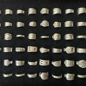 May include: A collection of silver rings displayed on a black surface. The rings vary in design, including plain bands, textured patterns, and floral motifs. The rings are arranged in neat rows, showcasing the variety of styles.
